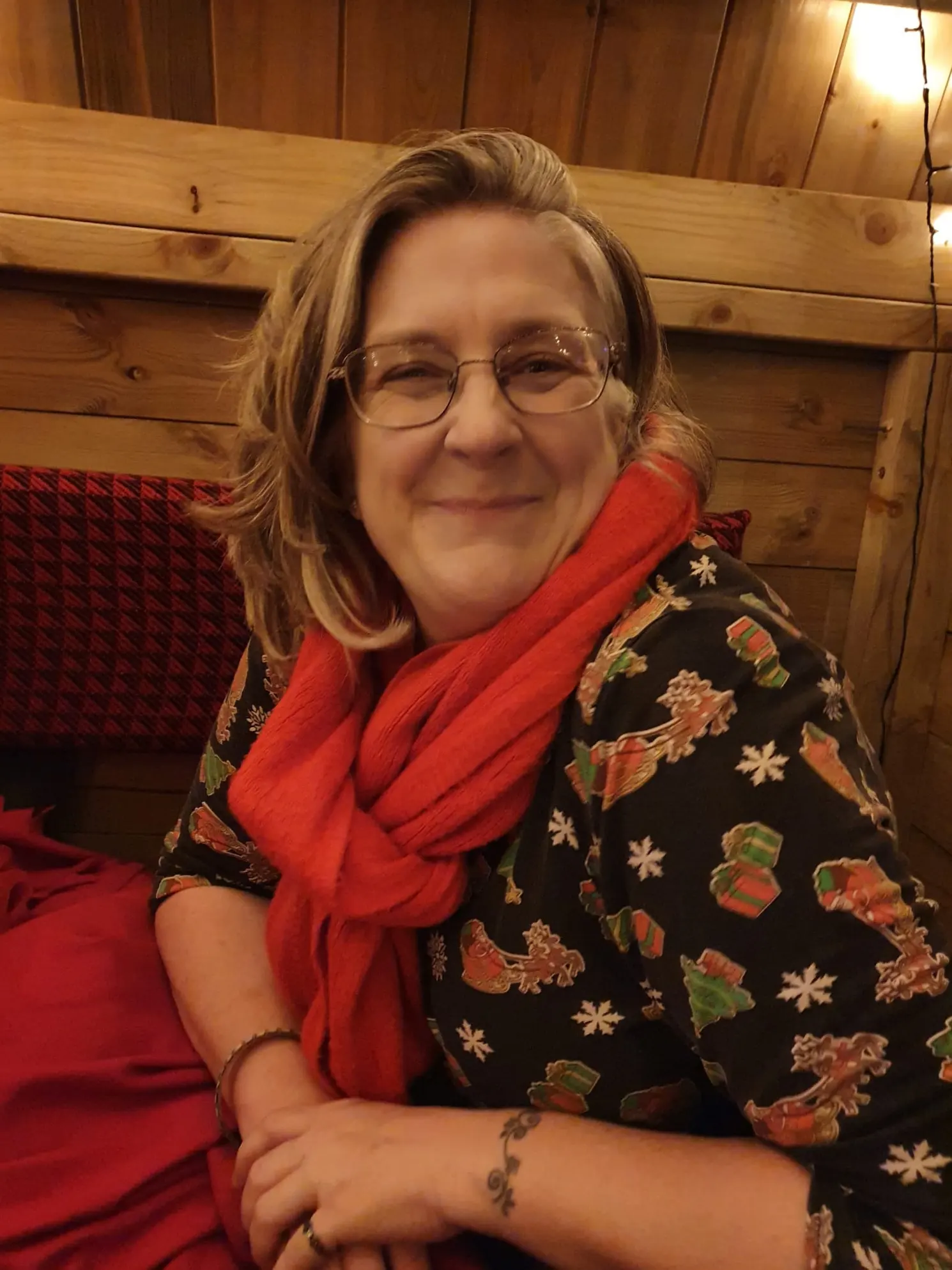 A woman smiling warmly at the camera while seated in a cozy wooden interior, wearing glasses, a festive black top with gift and snowflake prints, and a bright red scarf.